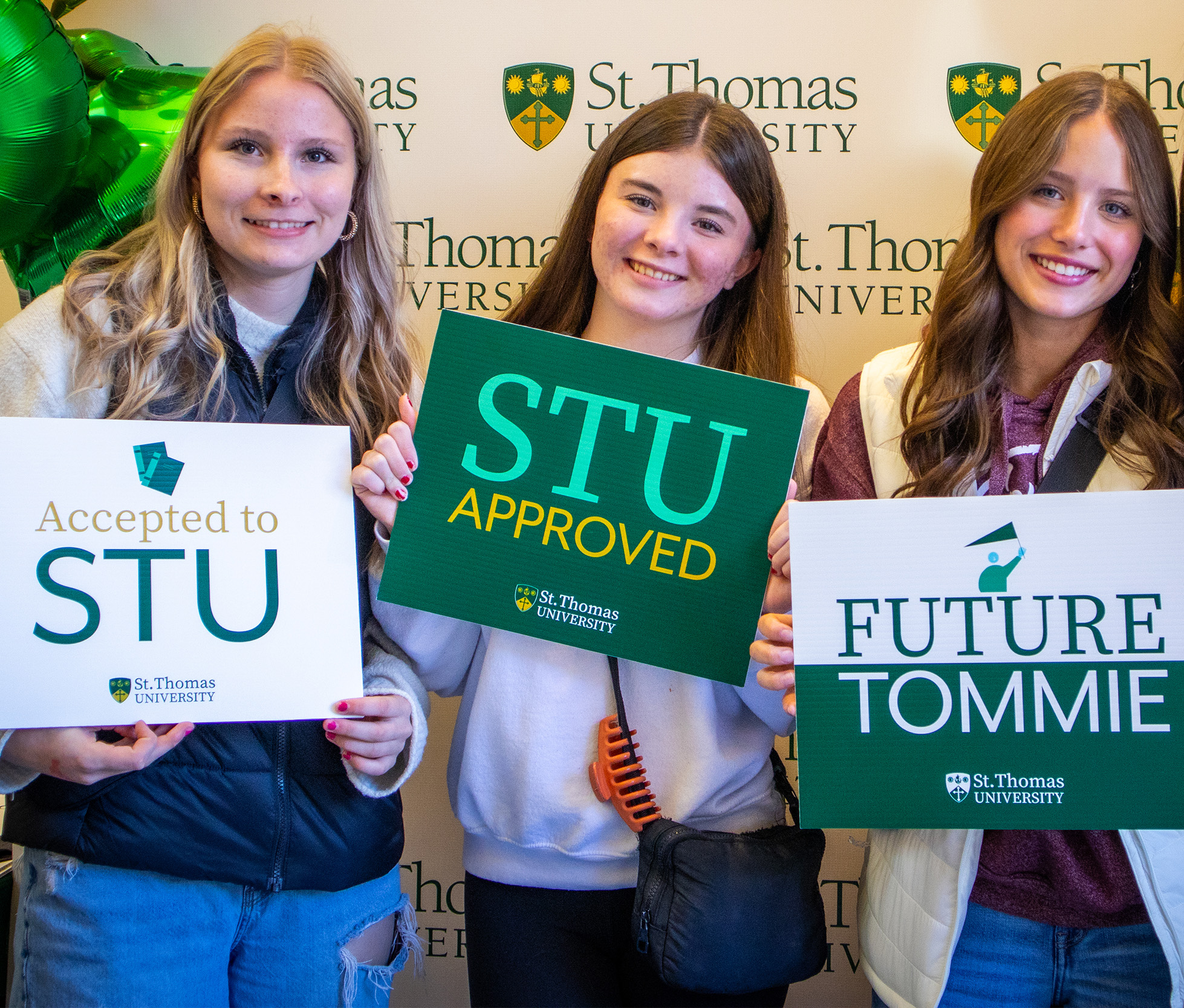 Three female students stand in front of a STU backdrop holding signs that say