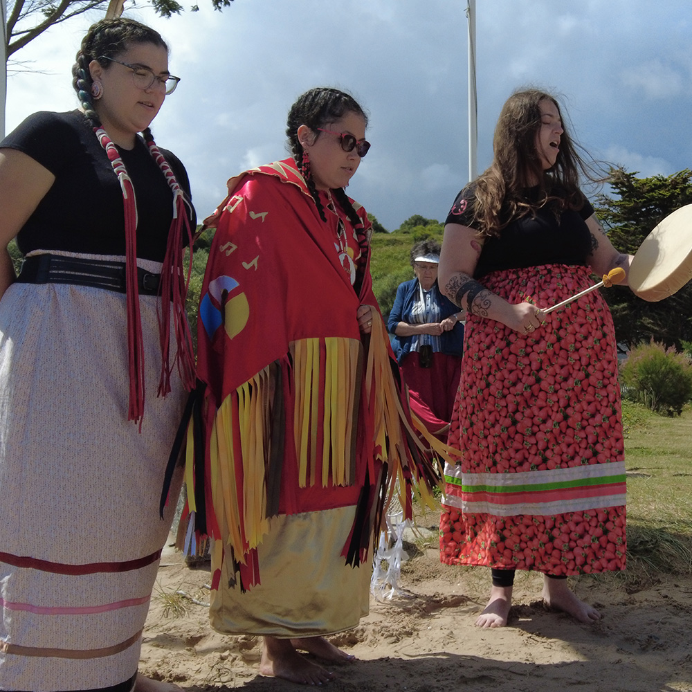Three Indigenous women wearing ribbon skirts stand outside singing