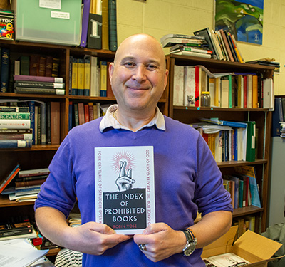 Dr. Robin Vose stands in his office holding up his latest book on the Index of Prohibited Books
